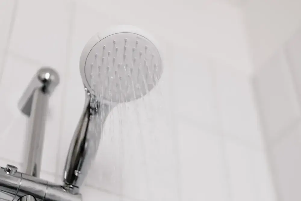 Pomme de douche d’où coule l’eau, devant une céramique blanche.
